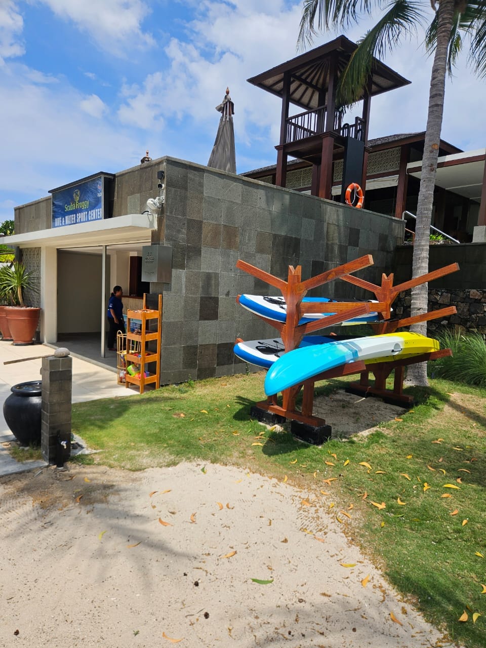 An outdoor, eye-level shot shows a Scuba Froggy diving and water sport center on a sunny day. A small, single-story building with grey tiled walls and a flat roof is in the center, with a blue sign above the entrance reading "Scuba Froggy DIVING & WATER SPORT CENTER". Inside, a person in a blue shirt is visible. To the right, a wooden rack holds stacked paddleboards and a kayak. In the background, a taller, dark wood structure with a lookout tower and a lifebuoy is visible. A palm tree stands tall behind the wooden structure. The ground is sandy with patches of grass and scattered dry leaves. The sky is blue with some clouds.
